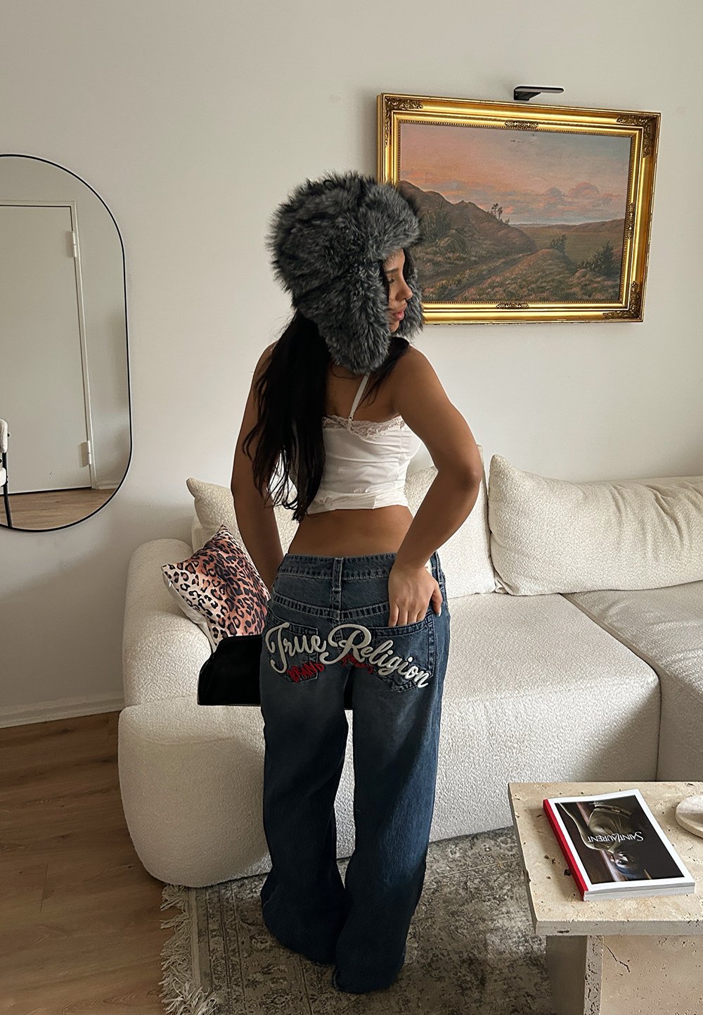 Jean denim à jambes larges avec "True Religion" brodé en blanc et rouge sur les poches arrière ; associé à un haut blanc orné de dentelle et un chapeau en fourrure.