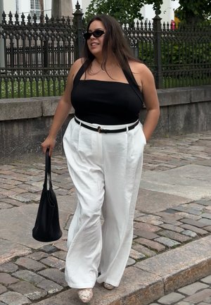 Top sin mangas negro, pantalones anchos blancos con un cinturón negro, llevando un bolso negro. Combinado con zapatos de diseño ligero. Fuera, sobre adoquines.