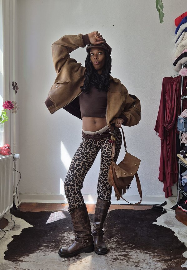 Femme posant à l'intérieur portant une veste courte marron, un pantalon imprimé léopard, des bottes marron et un sac à main et un chapeau assortis.