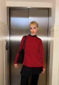 Jeune personne aux cheveux blonds courts portant une chemise rayée rouge et noire et une jupe plissée noire, debout devant des portes d'ascenseur fermées.