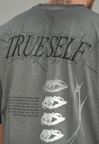 Grå skjorte bak med svart abstrakt tekst "TRUESELF" og sølv øye-grafikk ved siden av et motivasjonsavsnitt og abstrakt sølvdesign.