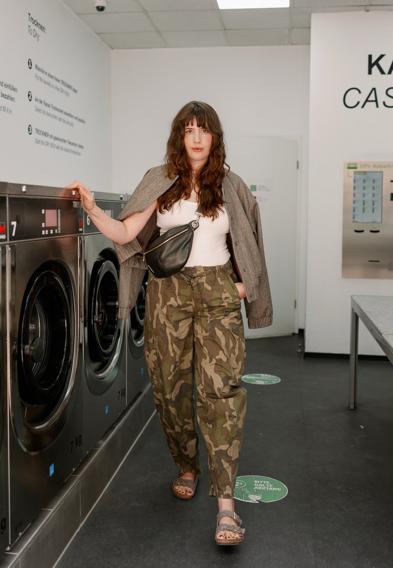 Femme portant un pantalon camouflage et une veste sur les épaules, debout dans une laverie, posant sa main sur une rangée de machines à laver à chargement frontal.