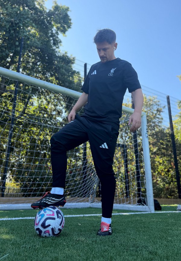 Hombre con ropa deportiva negra de pie sobre césped artificial con un pie sobre un balón de fútbol cerca de un poste de portería en un día soleado.