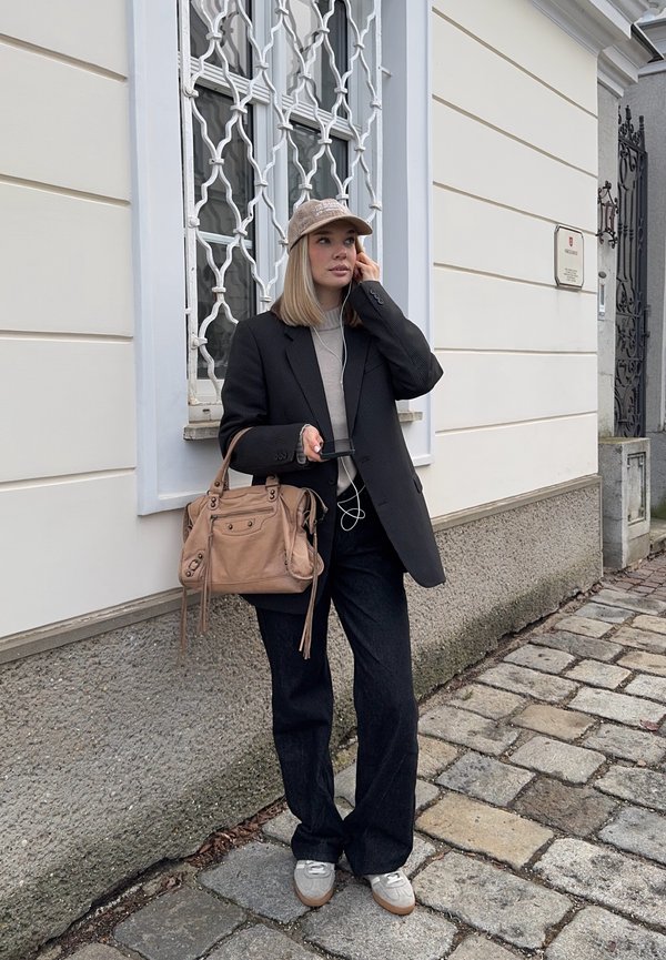 Junge Frau mit beiger Kappe, schwarzem Blazer und weiten Hosen, die eine beige Handtasche hält und auf einer Kopfsteinpflasterstraße Kopfhörer benutzt.