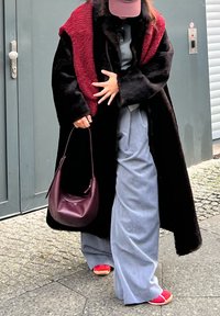 Nadrozmerný čierny kabát z umelej kožušiny s textúrovaným bordovým panelom, doplnený svetlomodrými širokými nohavicami, červenými topánkami a bordovou kabelkou na rameno.