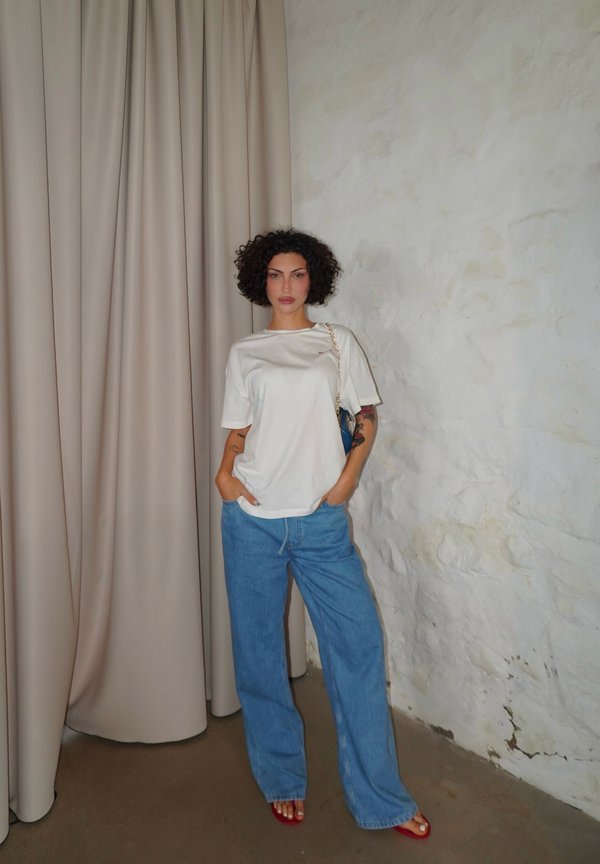 Woman with curly hair wearing a white t-shirt, blue wide-leg jeans, and red sandals standing against a white textured wall and beige curtain.