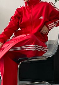 Rdeč Adidas trenirka z belimi črtami ob ramenskih delih in hlačah. Jakna na zadrgo, sijoča tkanina z gladko teksturo. Jasna ozadje s stolom.