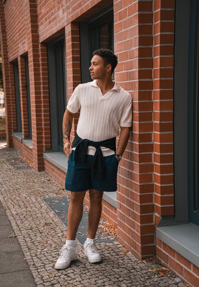 Joven con tatuajes, vistiendo una camiseta de punto blanca, pantalones cortos marinos y zapatillas blancas, apoyado contra un edificio de ladrillo en una acera.