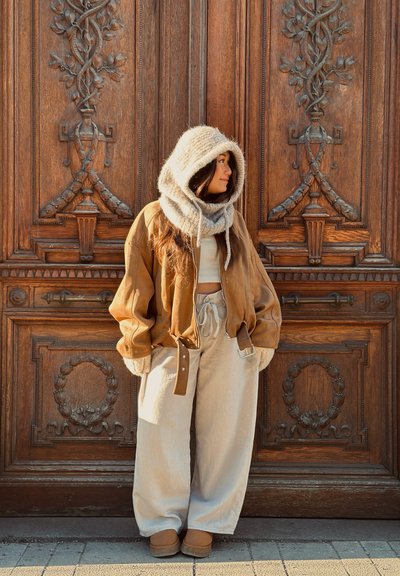 Mujer con una chaqueta marrón extragrande, pantalones anchos crema y bufanda de punto con capucha, de pie frente a una gran puerta de madera ornamentada, mirando hacia la derecha.