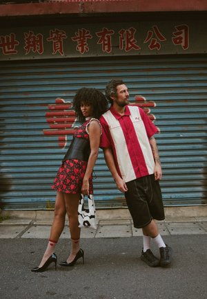 Vestido con estampado de corazones rojos, corsé de cuero negro, tacones negros. Hombre con camisa de manga corta roja y blanca, pantalones cortos negros y zapatillas.