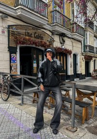 Person i svart hettejakke, baggy jeans og hodetelefoner som står på en gate nær en pub med utendørsbenker og sykler.