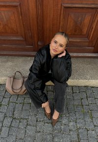 Młoda kobieta w czarnej kurtce, szarych dżinsach i butach w panterkę siedzi na kamiennym schodku obok drewnianych drzwi, a obok na ziemi leży beżowa torebka.