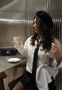 Femme portant un béret et une chemise rayée tenant une tasse blanche assise à une table en métal avec un sac noir, une assiette et une cuillère, devant un mur cannelé.