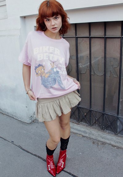 Mujer joven con cabello rojo que lleva una camiseta rosa de "Biker Betty", una falda beige con volantes, calcetas negras y botines rojos brillantes de tobillo, de pie sobre el pavimento.