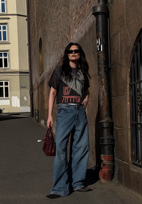 Femme portant un t-shirt Led Zeppelin, jean à jambes larges, chaussures pointues et lunettes de soleil, tenant un sac à main rouge, debout près d'un mur en briques et en pierre.