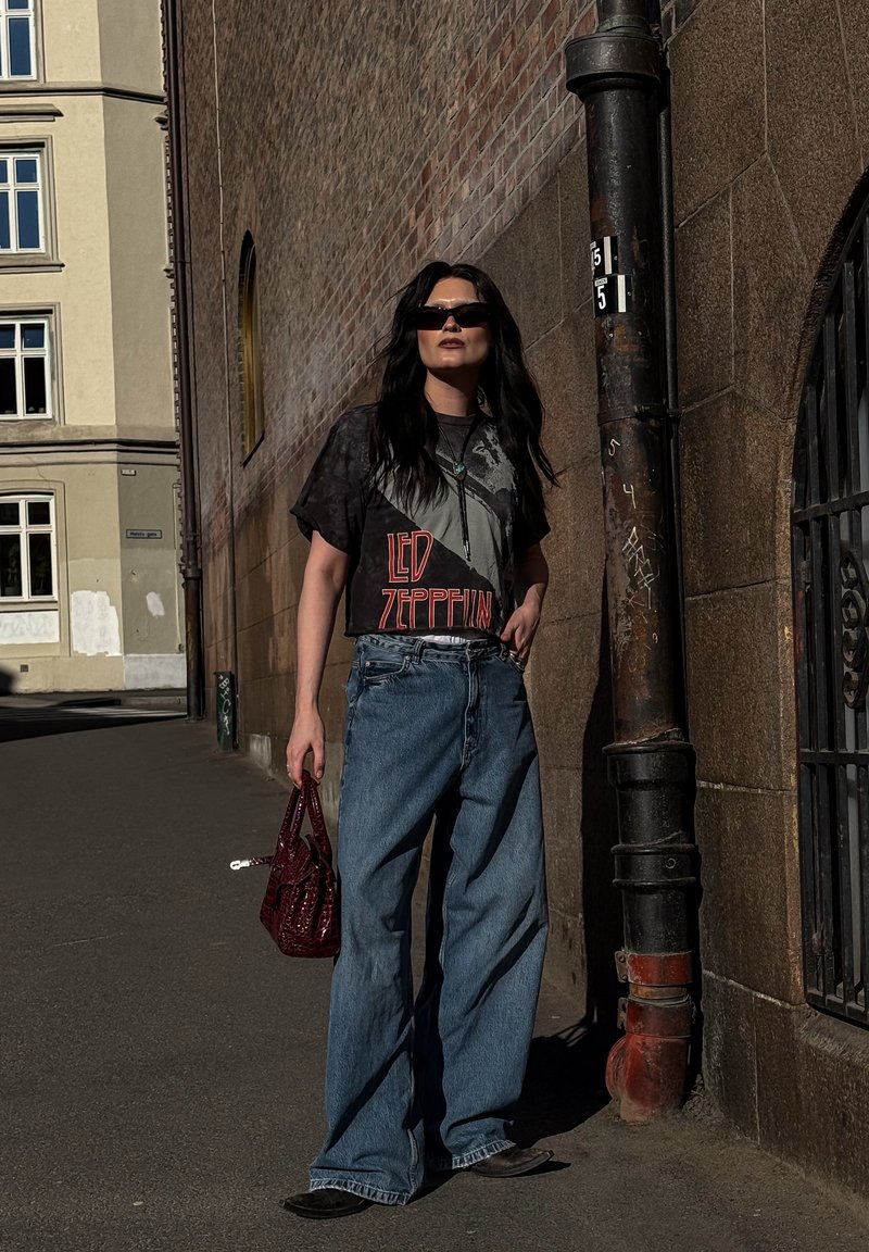 Žena so tričkom Led Zeppelin, širokými džínsami, špicatými topánkami a slnečnými okuliarmi, drží červenú kabelku, stojí pri tehlicovej a kamennej stene.