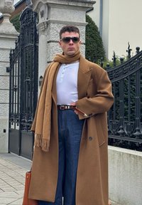 Homme portant un manteau camel, une écharpe assortie, une chemise henley blanche, un jean bleu, une ceinture marron et des lunettes de soleil, debout près de piliers en pierre et d'une clôture en fer.
