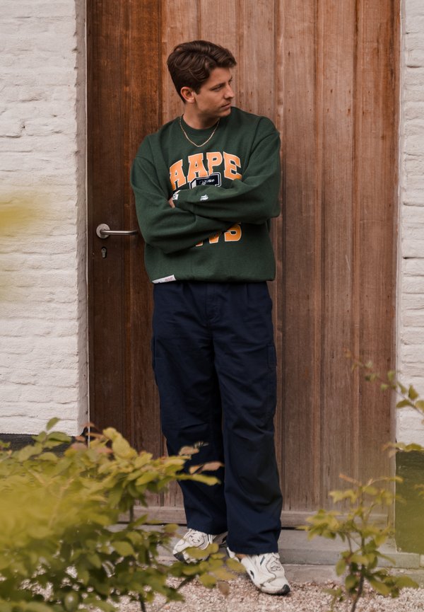 Sudadera verde con letras naranjas, pantalones cargo azul marino y zapatillas blancas. De pie junto a una puerta de madera, con los brazos cruzados, con follaje en primer plano.