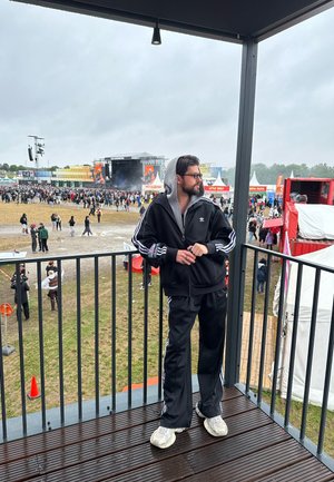 Schwarzer Adidas-Trainingsanzug mit weißen Streifen, grauer Hoodie, steht auf einem Balkon und blickt auf eine Menge bei einem Festival. Grauer Himmel im Hintergrund.