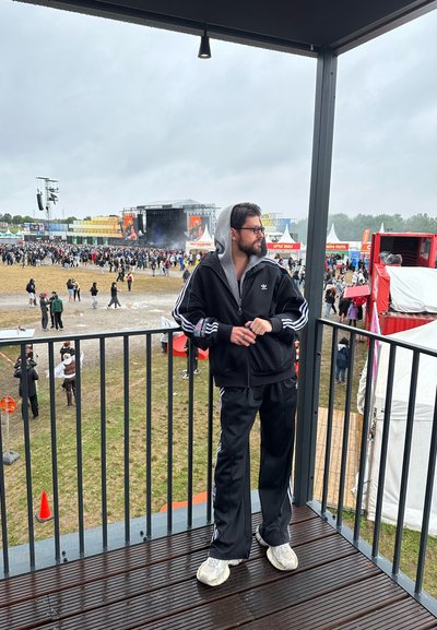Chándal negro de Adidas con rayas blancas, sudadera gris, de pie en un balcón con vistas a una multitud en un festival. Cielos grises de fondo.