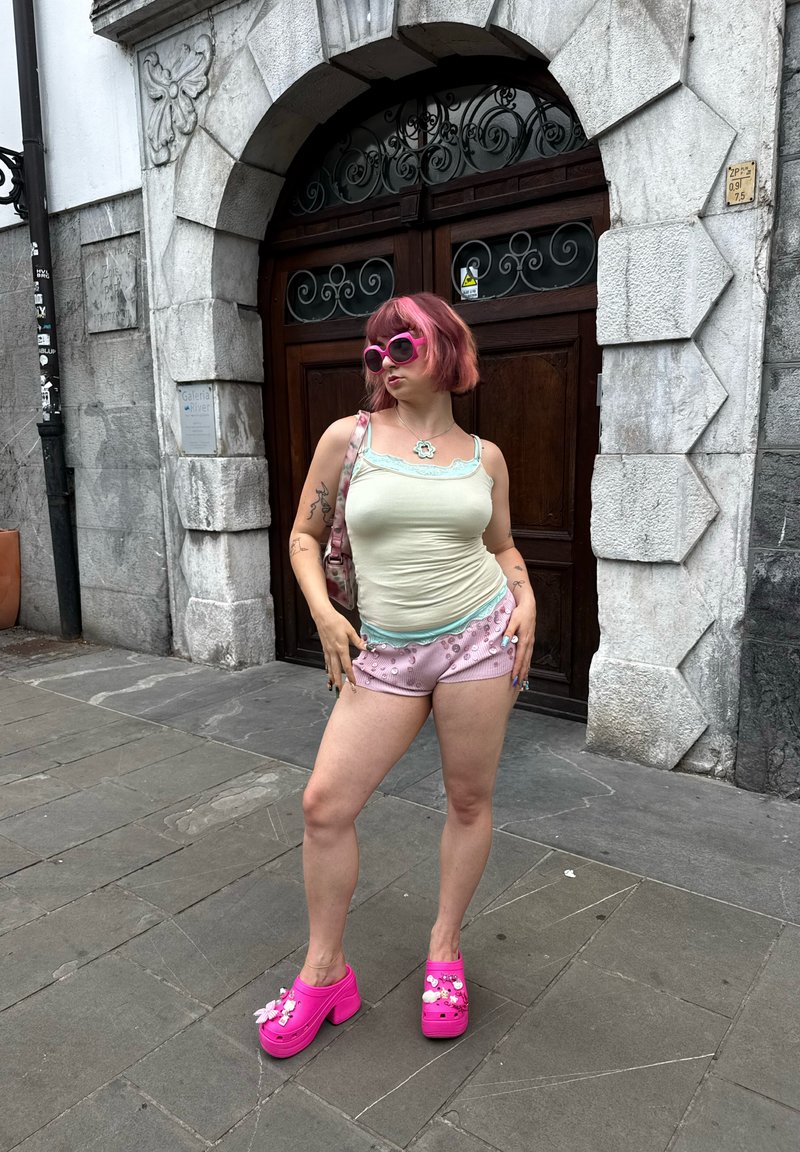 Femme aux cheveux roses, lunettes de soleil, short et chaussures compensées roses, debout avec assurance devant une porte en bois arquée avec encadrement en pierre.