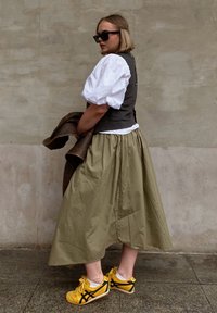 Λευκό πουκάμισο με φουσκωτά μανίκια, σκούρα γιλέκο και μακριά ελιά φούστα. Κίτρινες αθλητικές σκάφες με μαύρες λεπτομέρειες. Καφέ μπουφάν ριγμένο σε έναν αγκώνα.