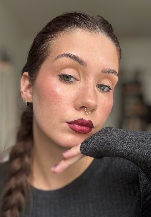 Portret van een persoon met lang bruin haar en een grijze trui, met opvallende bordeauxrode lippenstift en een neusring. Zonnesproetjes en groene ogen zichtbaar.