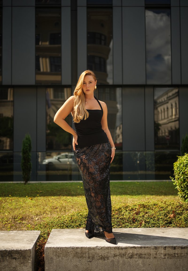 Mujer con cabello largo y rubio, vistiendo un top negro sin mangas y una falda negra de encaje transparente, de pie al aire libre frente a un edificio moderno de cristal.