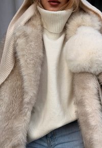 Femme portant un pull col roulé crème, un manteau en fausse fourrure beige avec capuche et un jean bleu, vue des épaules aux hanches.