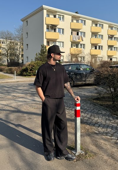 Camiseta negra oversize y pantalones negros holgados. Lleva una gorra negra y zapatillas deportivas. De pie junto a un poste plateado con detalles en rojo y blanco.