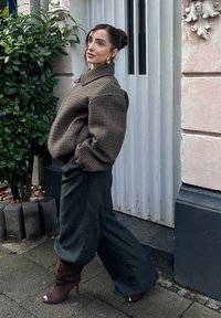 Braune übergroße Hahnentritt-Jacke, graue hochtaillierte Hose und braune geschnürte offene High Heels, vor einer strukturierten Wand und Grünpflanzen.