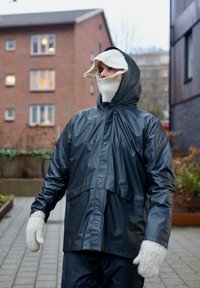 Czarna wodoodporna kurtka z kapturem, zapinana na zatrzaski, o luźnym kroju, w połączeniu z kremowymi, dzianymi rękawiczkami w fakturowanym wzorze.