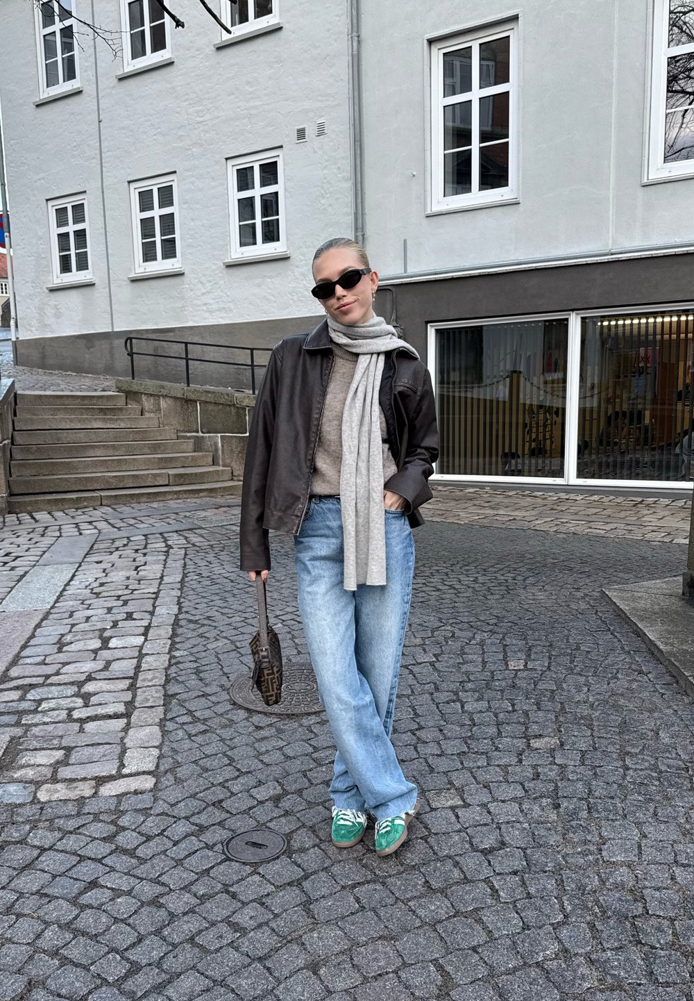 Vrouw met een zonnebril, leren jas, sjaal, losse jeans en groene sneakers staat op een straat met kinderkopjes voor een gebouw met ramen.