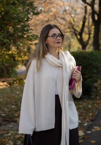 Bež, široki kardigan mekane teksture s otvorenim prednjim dijelom, ležerno prebačen. Glatka tkanina, uparena s tamnom suknjom; drži knjige.