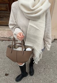 Personne portant un pull beige, une écharpe blanche, des bottes noires, tenant un sac à main marron avec de la fourrure blanche et une tasse de café à l'intérieur.