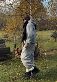 Set di felpa e pantaloni grigio chiaro, realizzato in un tessuto morbido. Una sciarpa nera valorizza l'outfit. Stivaletti neri, ambiente naturale con fogliame autunnale.