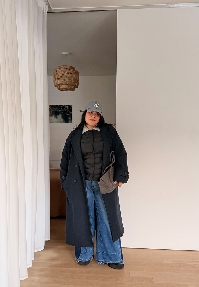 Femme portant un long manteau noir, une doudoune noire, un jean à jambes larges bleu, une casquette de baseball grise NY, et tenant un sac à main gris à l'intérieur.