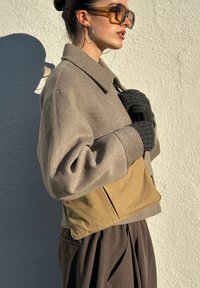 Veste marron coupée courte avec un col, présentant un panneau inférieur beige, un tissu texturé, et assortie à des gants en tricot noirs.