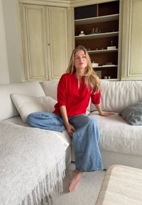 Mujer con cabello rubio largo que lleva una blusa roja y jeans de mezclilla anchos, sentada descalza en un sofá seccional beige en una sala de estar.