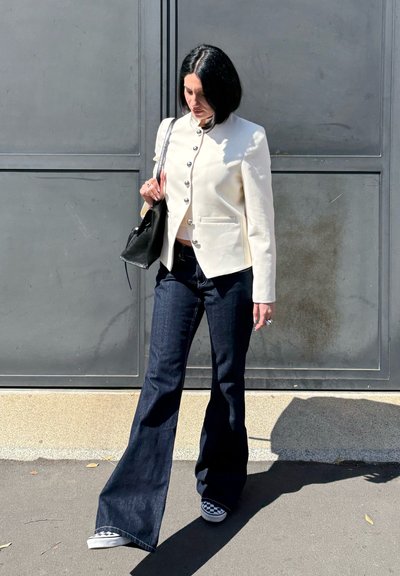 Mujer con cabello corto y negro lleva una chaqueta blanca abotonada, vaqueros acampanados oscuros, zapatos tipo slip-on con patrón de tablero de ajedrez y lleva un bolso negro al aire libre.