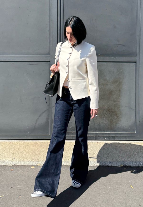 Femme aux cheveux noirs et courts porte une veste blanche boutonnée, un jean évasé foncé, des chaussures slip-on à motif damier et porte un sac à main noir en extérieur.