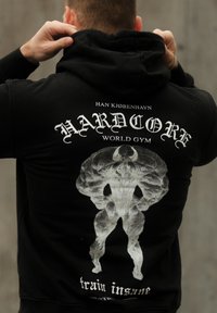 A person adjusting a black hoodie featuring a white graphic of a muscular figure along with the text "HARDCORE World Gym, train insane" on the back.
