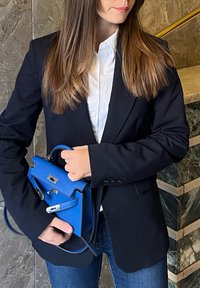 Borsa blu in pelle texturizzata, con manico superiore, hardware argentato e chiusura a pattina. Indossata con un blazer nero e una camicia bianca.