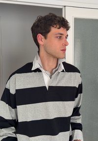 Jeune homme aux cheveux bouclés et foncés portant un maillot de rugby rayé noir et gris, debout à l'intérieur, regardant vers la droite.