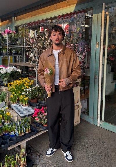 Chaqueta de cuero marrón, camiseta blanca y pantalones anchos negros. Sosteniendo un ramo de tulipanes rosas en papel kraft, rodeada de flores.
