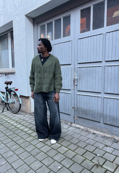 Cardigan verde de punto, sobredimensionado, abotonado en la parte delantera, llevado sobre una camiseta negra. Unos vaqueros oscuros de pierna ancha y unas zapatillas blancas completan el conjunto.
