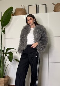 Jeune femme portant une veste grise pelucheuse, une chemise blanche et un pantalon de survêtement noir Adidas, debout à l'intérieur à côté d'une plante verte et de placards blancs.