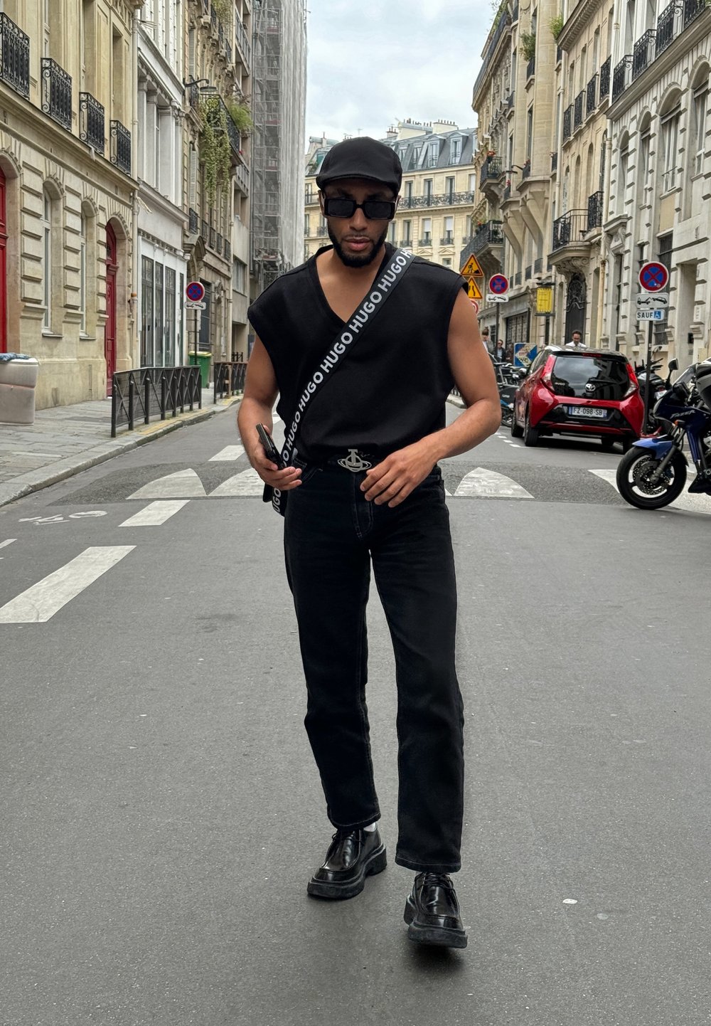 Μαύρο αμάνικο μπλουζάκι με λουρί με λογότυπο, σκούρα τζιν και γυαλιστερές μαύρες παπούτσια. Αστική σκηνή με κτήρια και οχήματα στο παρασκήνιο.