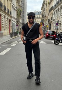 Μαύρο αμάνικο μπλουζάκι με λουρί με λογότυπο, σκούρα τζιν και γυαλιστερές μαύρες παπούτσια. Αστική σκηνή με κτήρια και οχήματα στο παρασκήνιο.