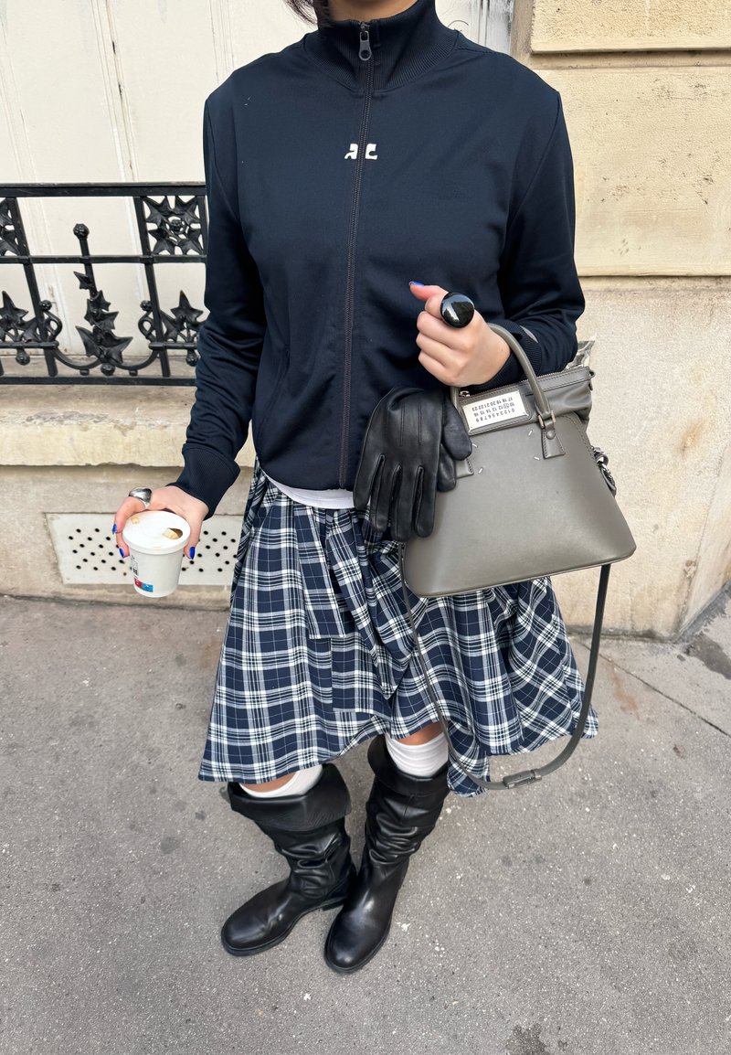 Personne portant une veste noire zippée, une jupe à carreaux et des bottes noires tenant un sac à main gris avec des gants et un gobelet de café jetable.